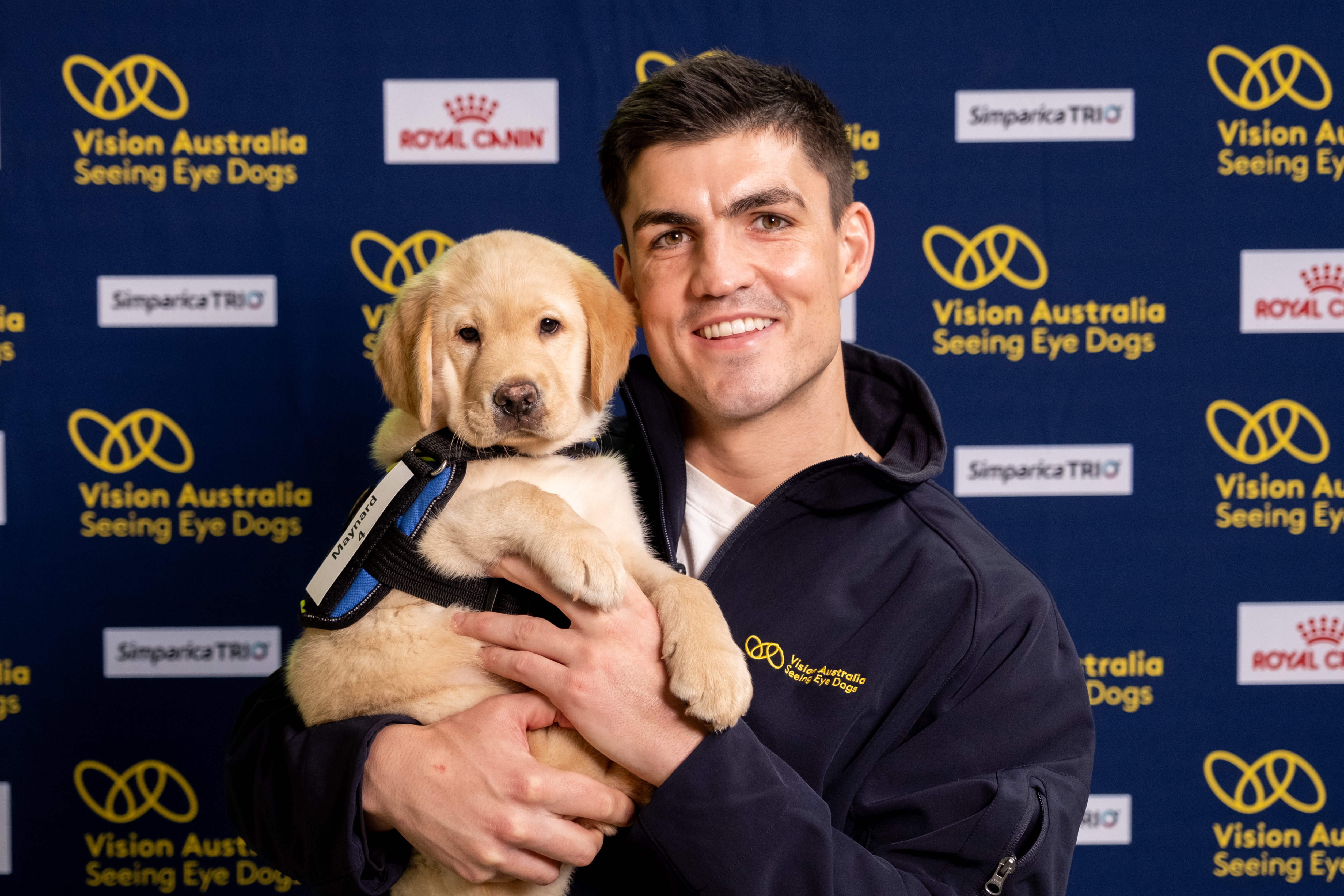 "Brayden Maynard smiling and holding a yellow Seeing Eye Dogs puppy in jacket in front of a Seeing Eye Dogs branded media wall."