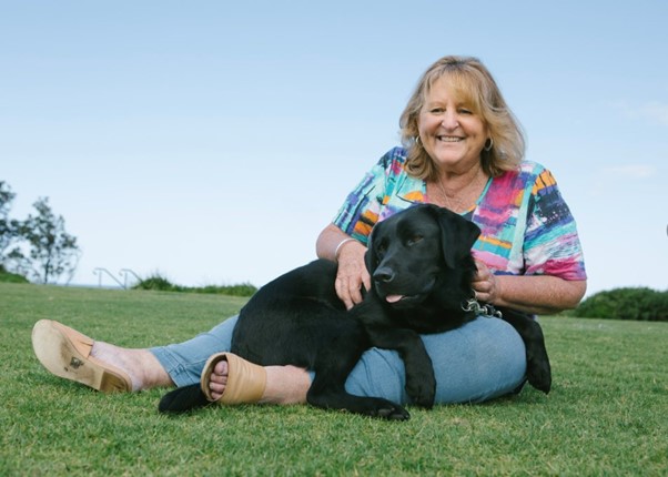 Cassie is reclined on the ground, with her Seeing Eye Dog Neesha sitting on her lap