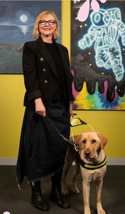 A lady in black clothes in standing, holding onto the lead of her Seeing Eye Dogg.