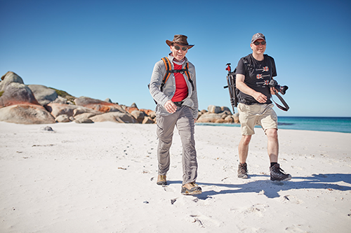 Two men walking along the coastline both wearing backpacks and one is holding a camera