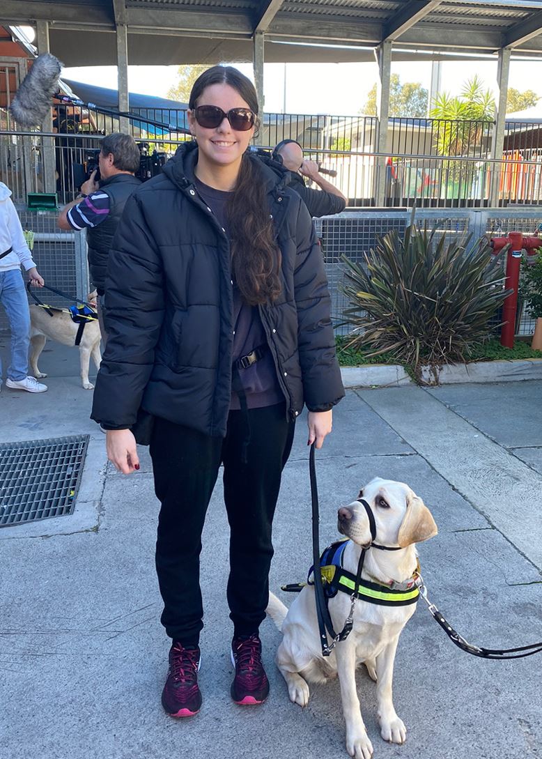 A girl wearing a dark puffer and sunnies smiles at the camera while holding the lead of a yellow Seeing Eye Dog wearing a harness